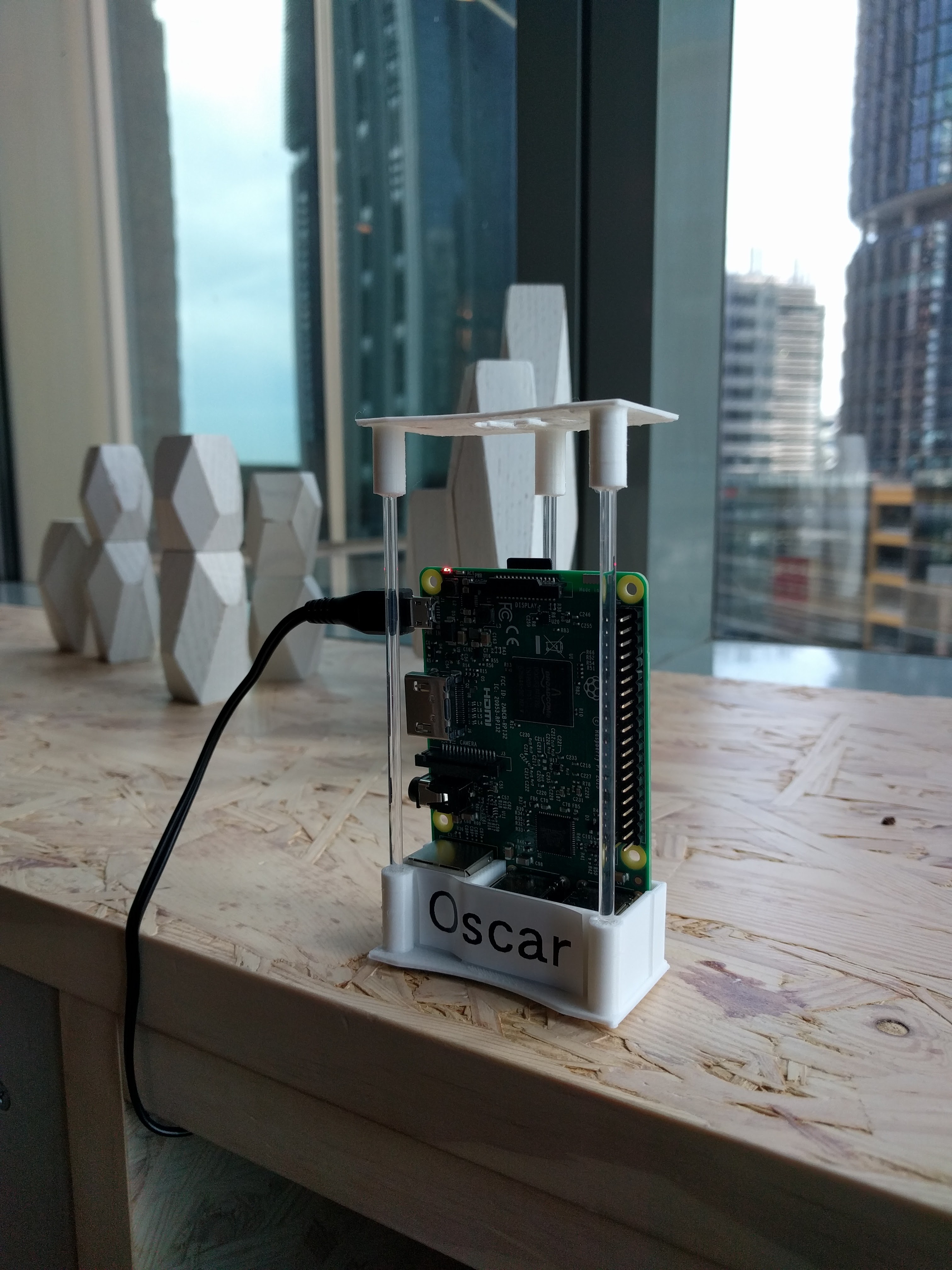A Raspberry Pi agent named "Oscar" in a clear enclosure, mounted by an office window with the city skyline behind it