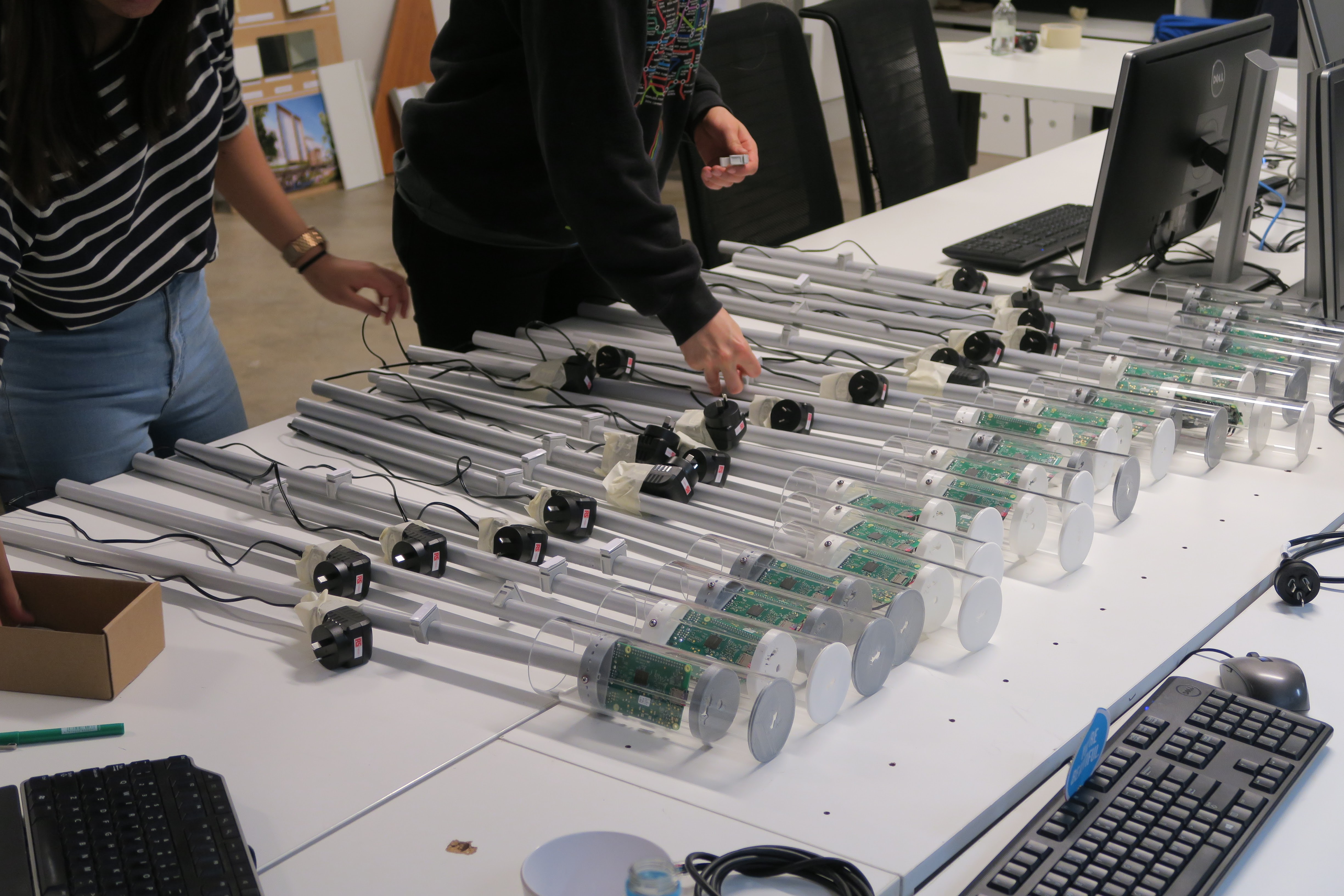 An assembly line of agents on an office desk: over a dozen clear acrylic tubes with Raspberry Pis inside, power plugs attached, being put together by two team members